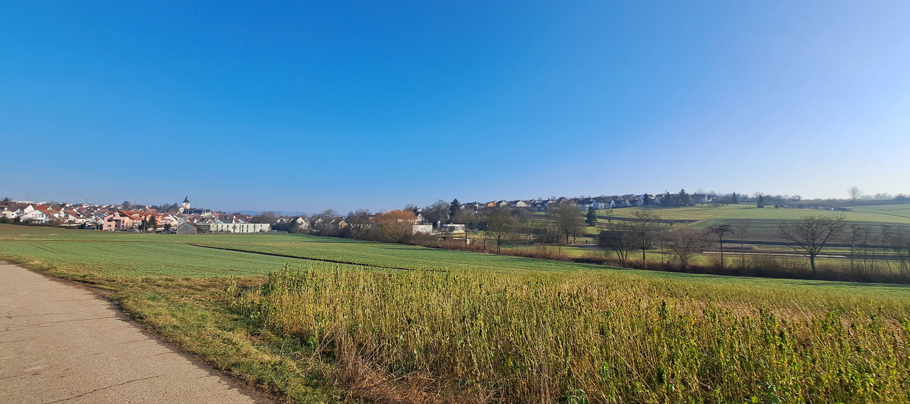 Weitblick über Felder und Wiesen auf die Ortschaft und Häusern unter klarem blauem Himmel.