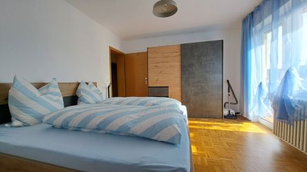 Schlafzimmer mit Doppelbett, großem Kleiderschrank und bodentiefem Fenster mit blauen Vorhängen.