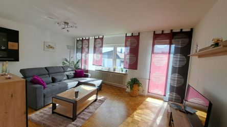 Helles Wohnzimmer mit grauer Couch, modernem Holztisch und großen Fenstern mit roten und grauen Vorhängen.